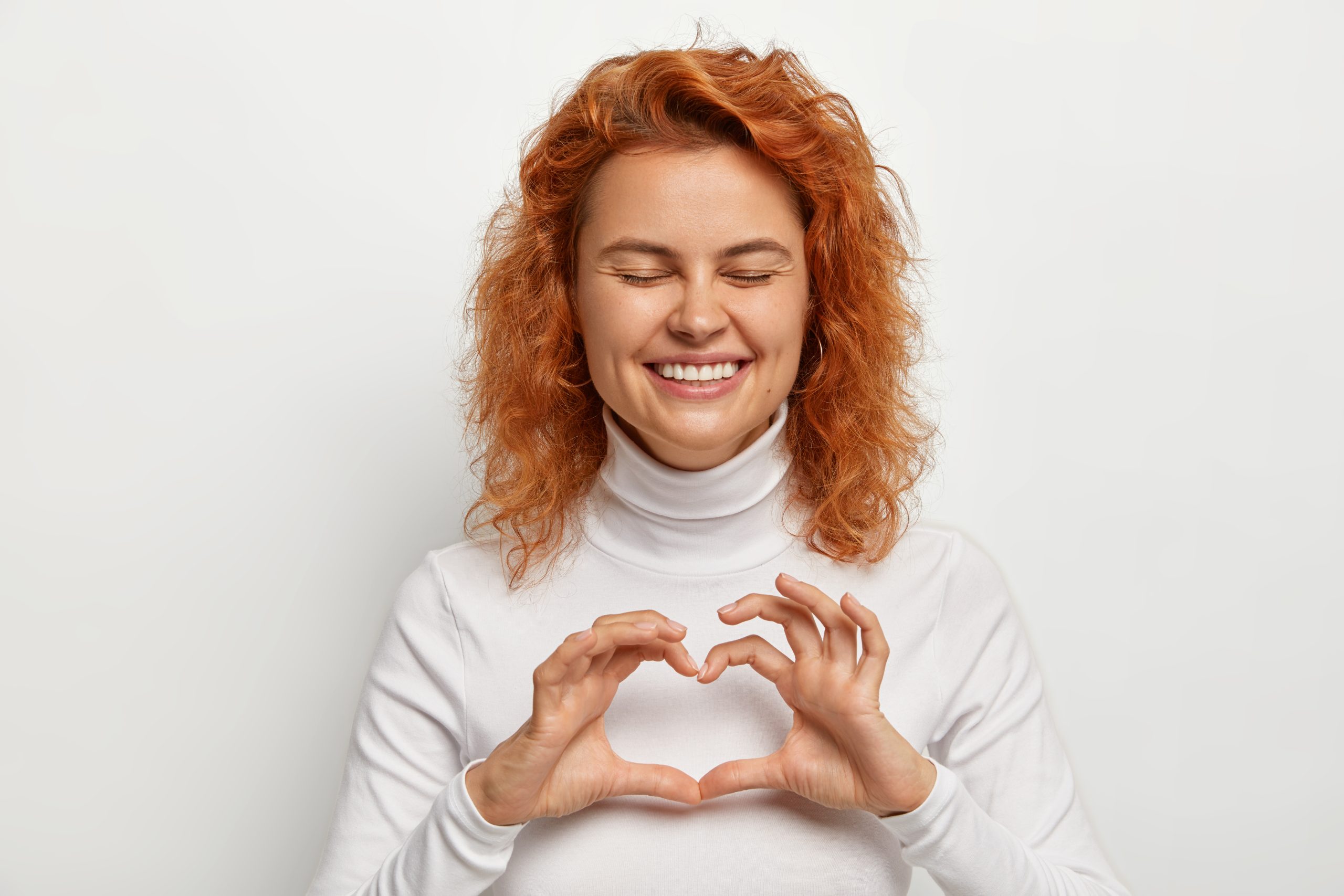 overjoyed redhead curly woman laughs positively shares love with you makes heart sign with hands chest expresses affection keeps eyes shut from pleasure dressed white casual outfit 300x200 1